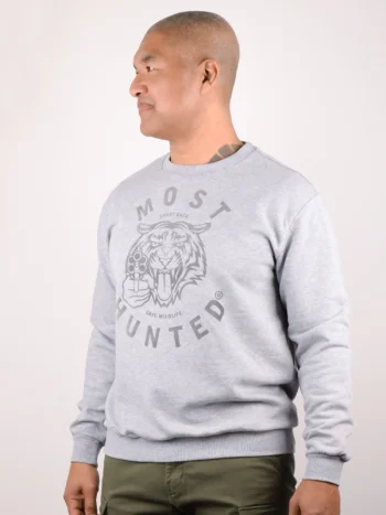 Side view of a male model looking away from the camera, wearing the grey melange MOST HUNTED unisex sweater with the large grey flock print of the tiger head and the texts 