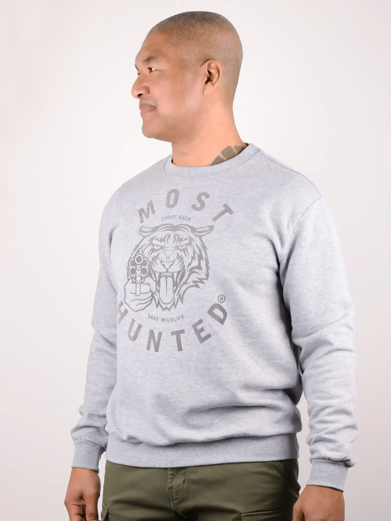 Zijaanzicht van een mannelijk model dat van de camera wegkijkt, die de grijs gemêleerde MOST HUNTED unisex sweater draagt met de grote grijze flockprint van de tijgerkop en met de teksten 